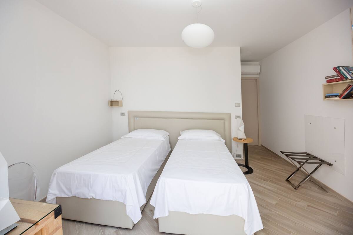 A minimalist bedroom with two single beds, a bedside table, a hanging light, and a bookshelf.