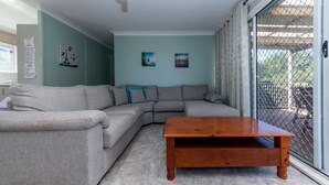 A living room with a grey sectional sofa, a wooden coffee table, and a sliding glass door leading to an outdoor area. - Right across the road from Fingal Bay Surf Beach (Fingal Bay)