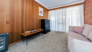 A living room with wooden panel walls, a black dresser, a beige sofa, and a small table with a laptop and a basket. - Right across the road from Fingal Bay Surf Beach (Fingal Bay)