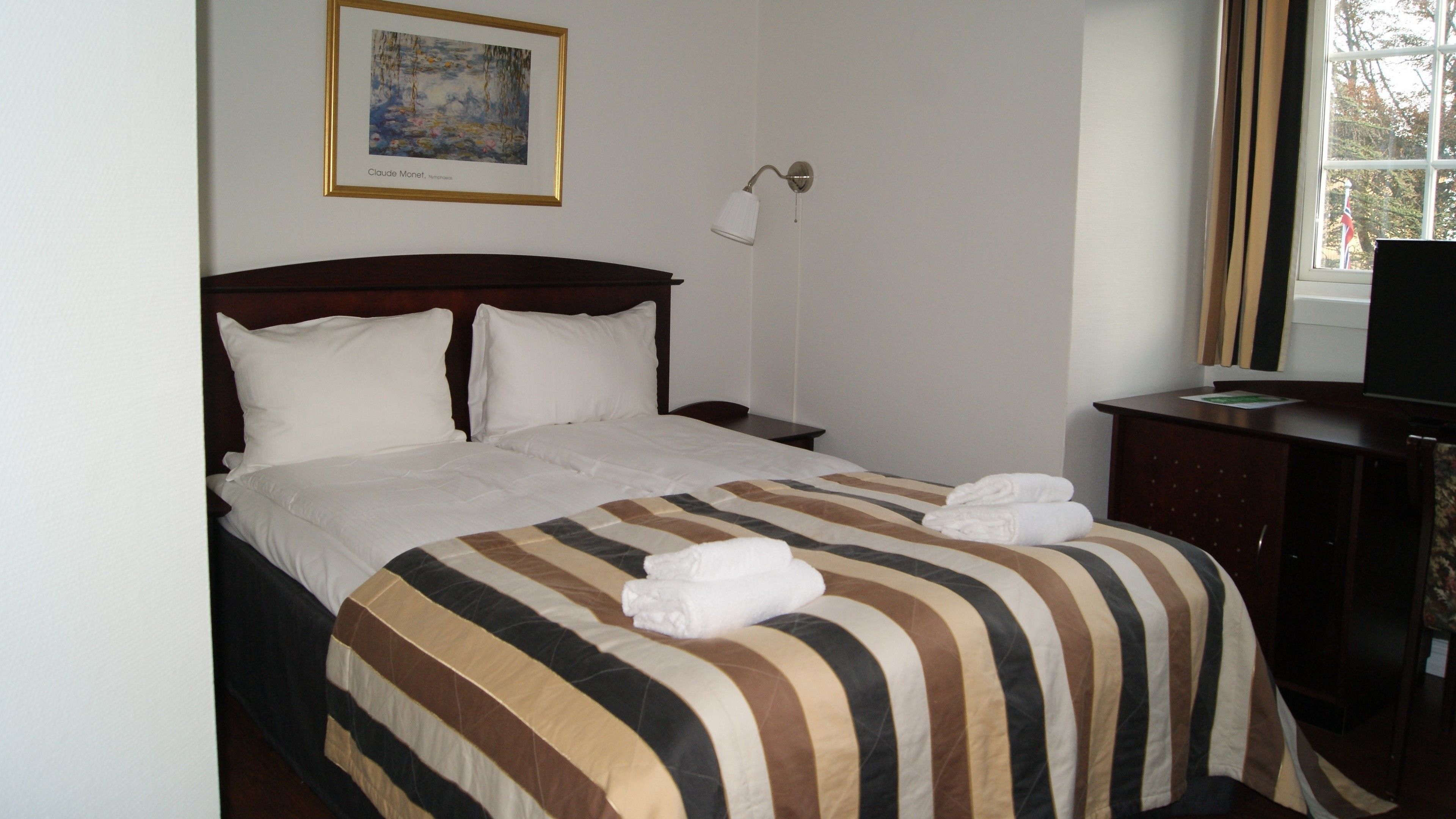 Habitación de hotel con cama, mesita de noche, lámpara, escritorio con televisor y ventana con cortinas.