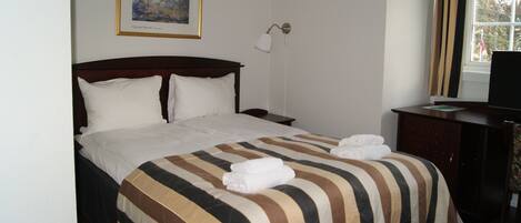 Habitación de hotel con cama, mesita de noche, lámpara, escritorio con televisor y ventana con cortinas.