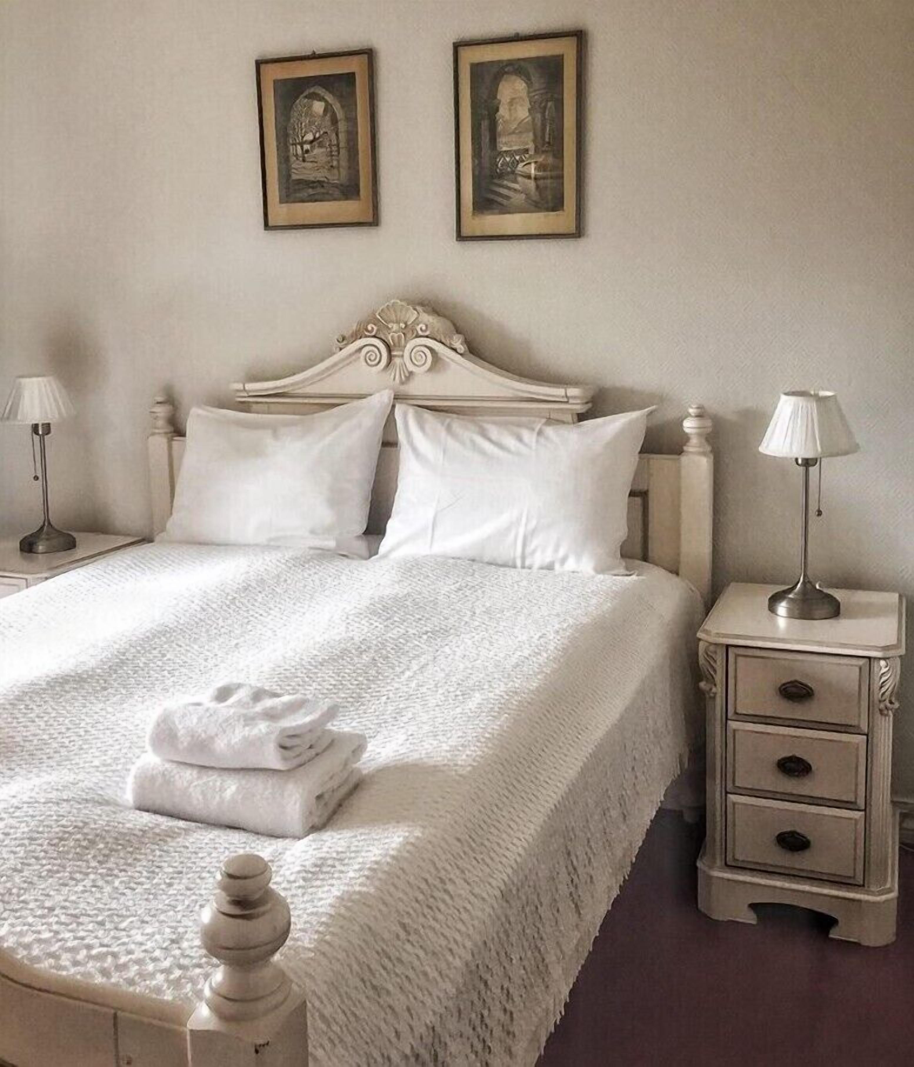A neatly made bed with white linens and pillows, a bedside table with a lamp, and two framed pictures on the wall.