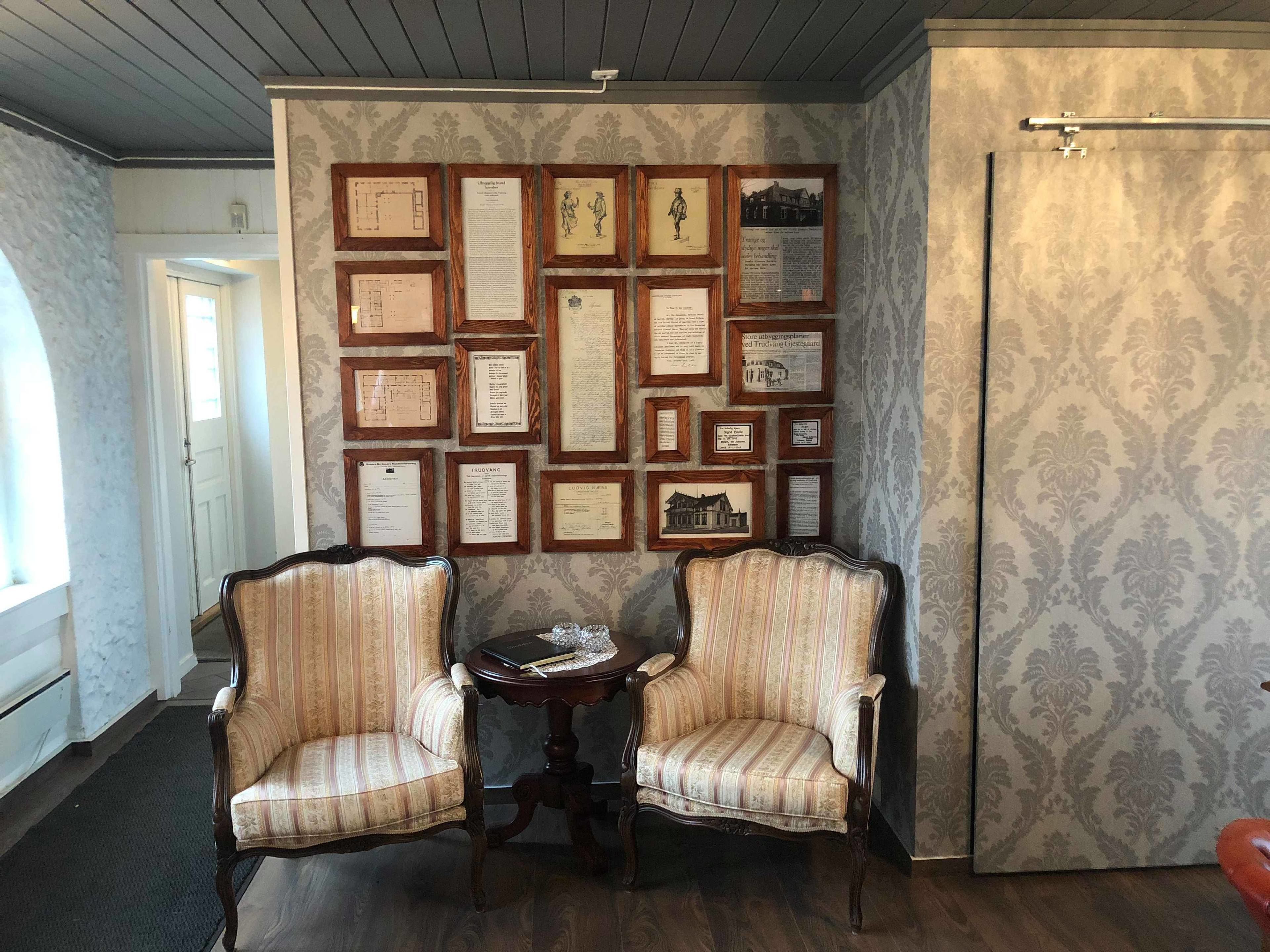 Una habitación con dos sillones tapizados, una mesita redonda y una pared decorada con documentos enmarcados.