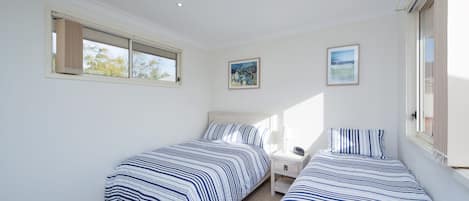 Un dormitorio con dos camas, una mesita de noche, un ventanal con cortinas y dos cuadros en la pared.