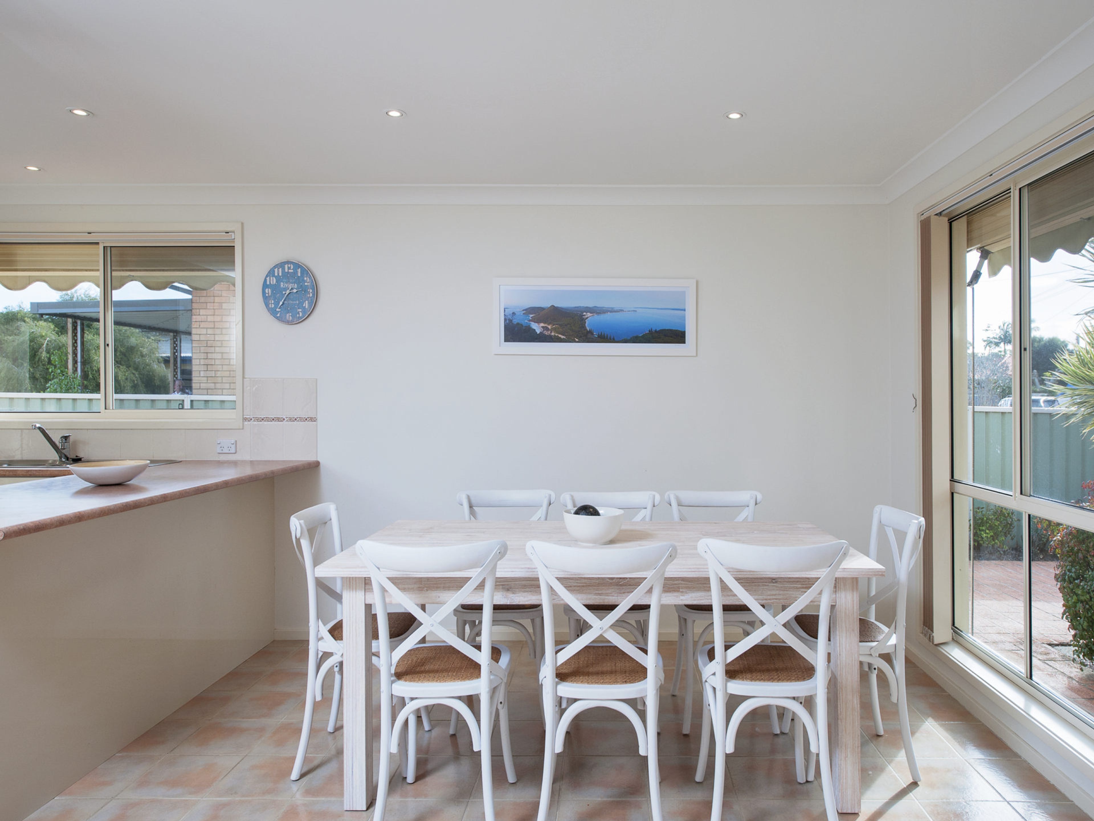 A dining area with a table and chairs, a clock on the wall, and a window with a view.