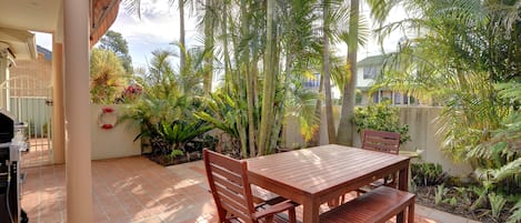 An outdoor patio with a wooden table and chairs, surrounded by lush tropical plants and a brick floor.