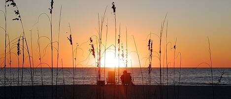 A sunset over a beach with tall grasses in the foreground and a chair facing the sunset.