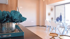 Una cocina moderna con una planta verde azulada en la encimera, un armario de madera y un comedor con sillas blancas y una mesa de vidrio.