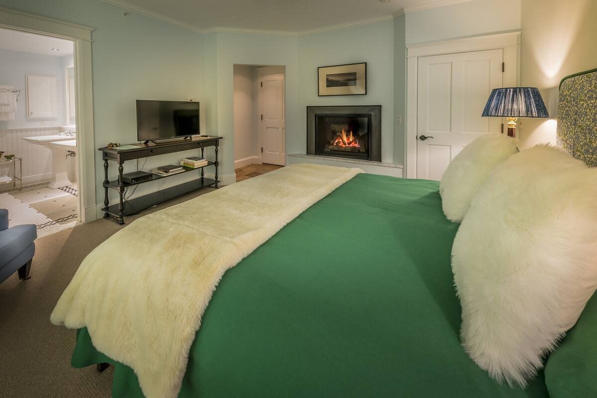 A bedroom with a green bedspread, a white fluffy pillow, a fireplace, a television on a stand, and a door leading to another room.