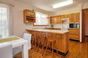 Een functionele keuken met houten kastjes, een centraal kookeiland met krukken, een wit aanrechtblad en een eethoek met een witte tafel en stoelen.
