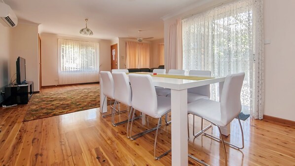 A modern living room with a dining area, wooden flooring, and large windows with sheer curtains.