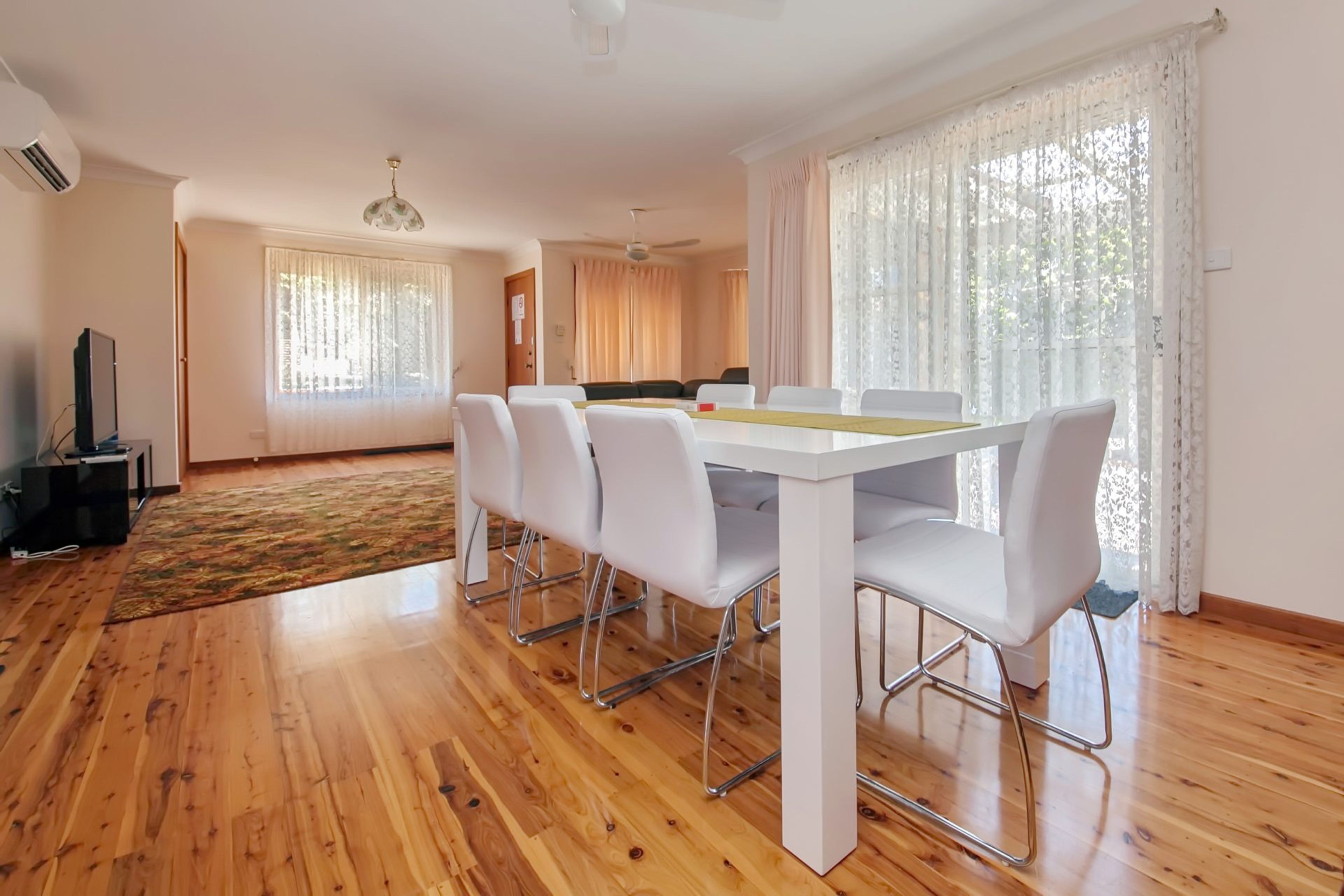 A modern living room with a dining area, wooden flooring, and large windows with sheer curtains.