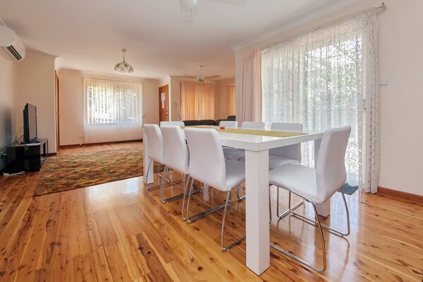 A modern living room with a dining area, wooden flooring, and large windows with sheer curtains.
