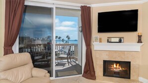 A living room with a beige sofa, a fireplace, a TV mounted on the wall, and a sliding glass door leading to a balcony with a view of the beach. - Pier View Escape (Oceanside)