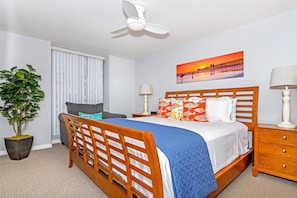 A bedroom with a wooden bed, a nightstand, a lamp, a potted plant, a window with blinds, and a sunset painting on the wall. - Our Ocean View (Oceanside)