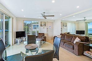 A spacious living room with a glass dining table, comfortable seating, a flat-screen TV, and a ceiling fan.