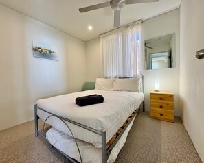 Un dormitorio con una cama individual, un aparador de madera, un ventilador de techo y una ventana con cortinas traslúcidas.