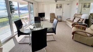 Een moderne woonkamer met een glazen tafel, comfortabele zitmogelijkheden, een flatscreen televisie en uitzicht op een meer.