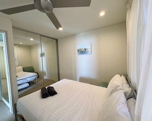 A hotel room with a bed, a ceiling fan, a mirror, a painting, and a window with curtains.