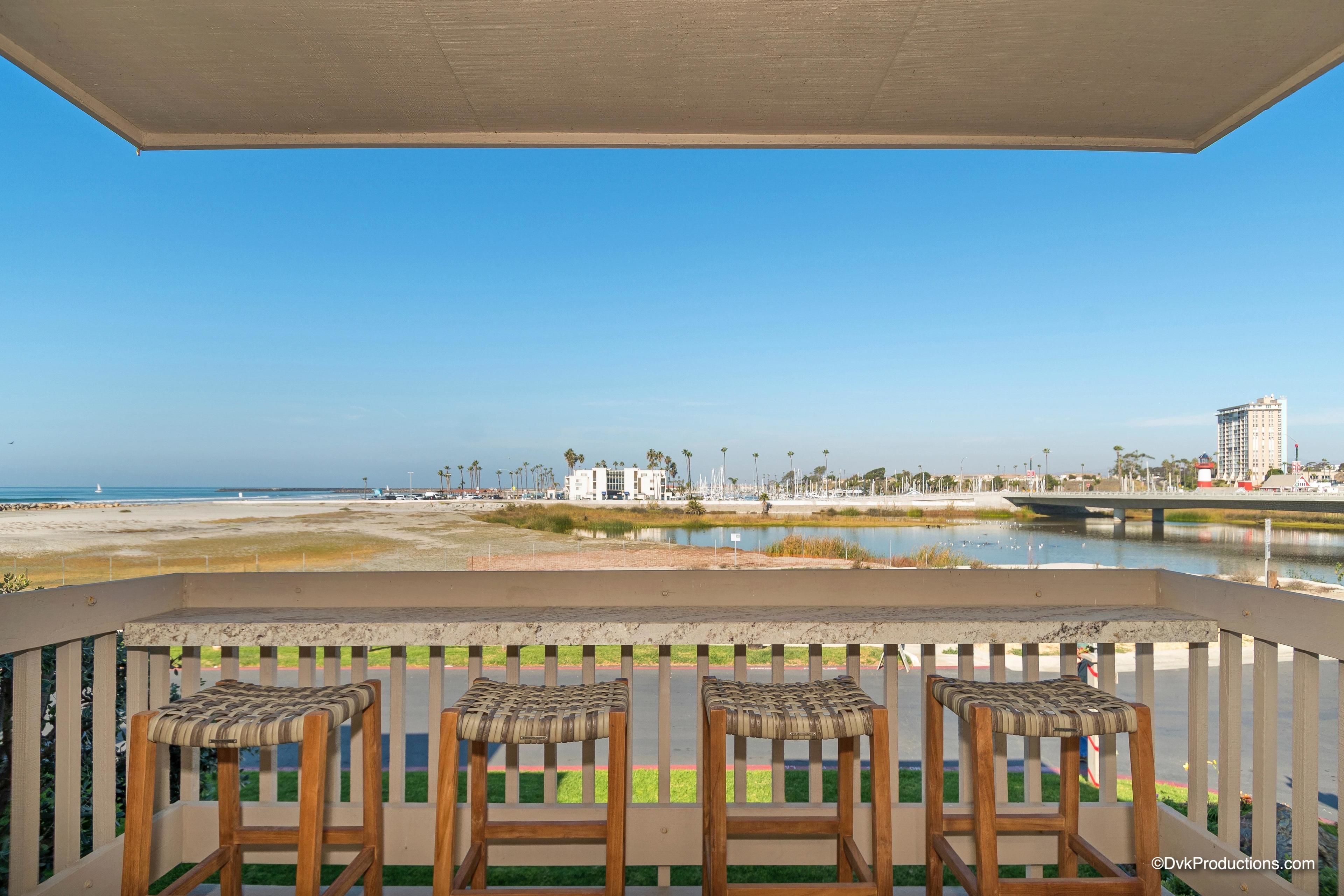 Un balcón con tres sillas de mimbre, vista a la playa y un paisaje urbano de fondo.