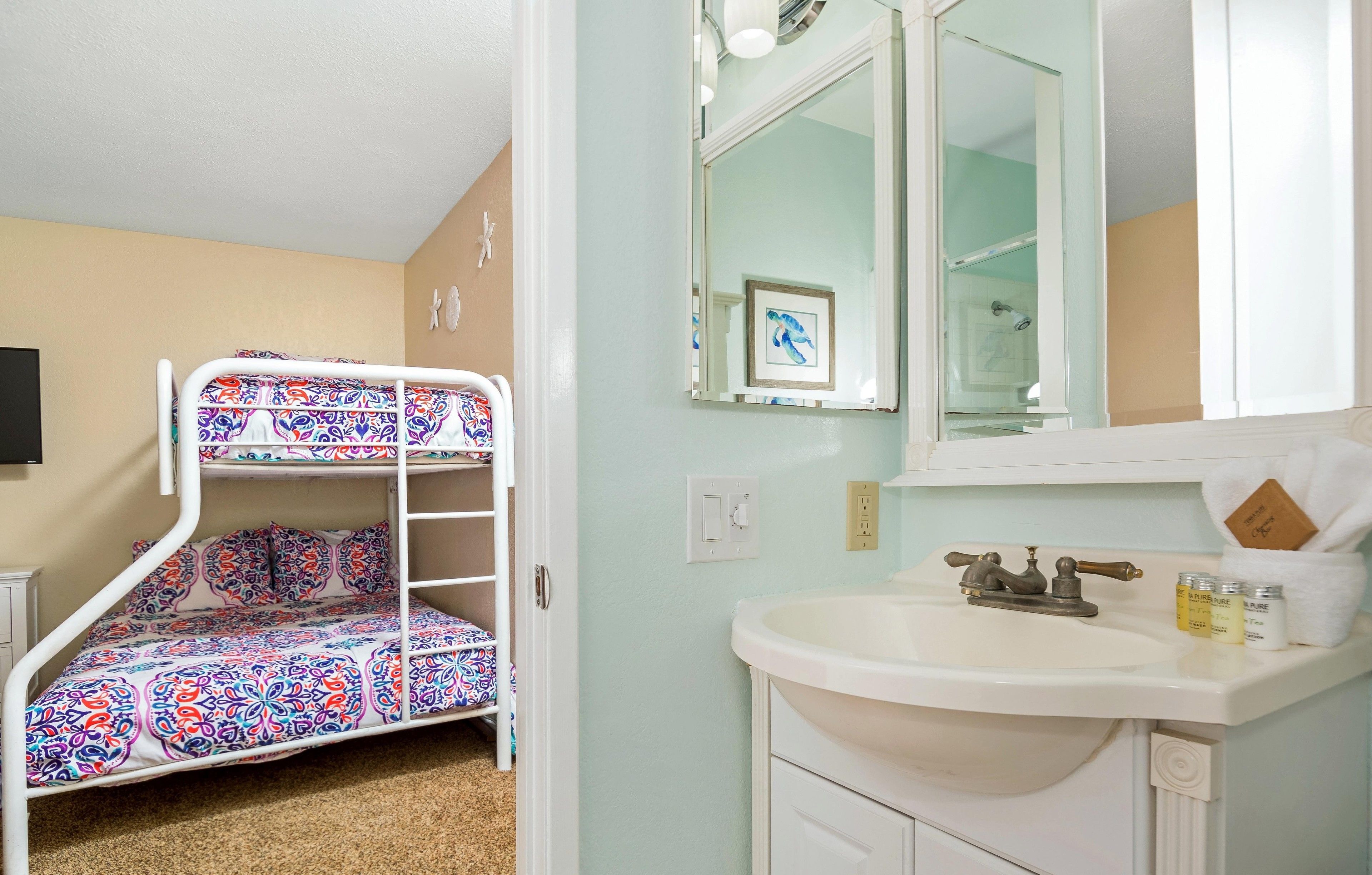 A bunk bed with a colorful patterned bedspread, a white sink with a mirror above it, and a framed picture on the wall.