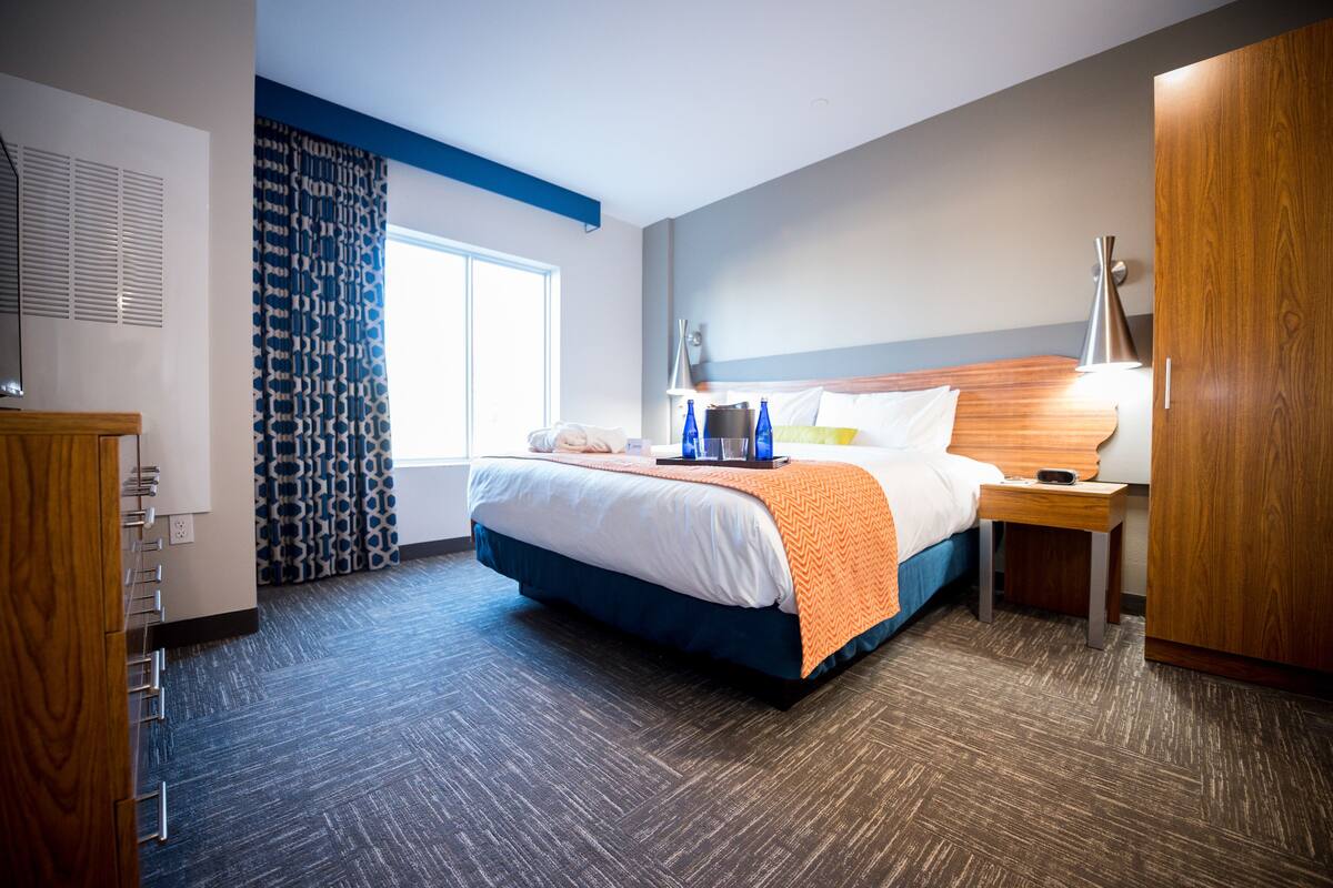 Habitación de hotel con una cama grande, una mesita de noche con lámpara, un armario y una ventana con cortinas estampadas.