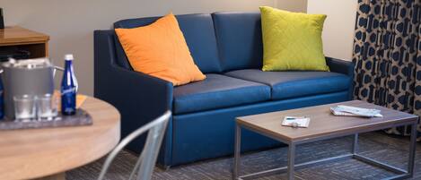 Una habitación de hotel moderna con un sofá azul, dos almohadas, una mesa de centro de madera, una silla y una mesita con una tetera y botellas.