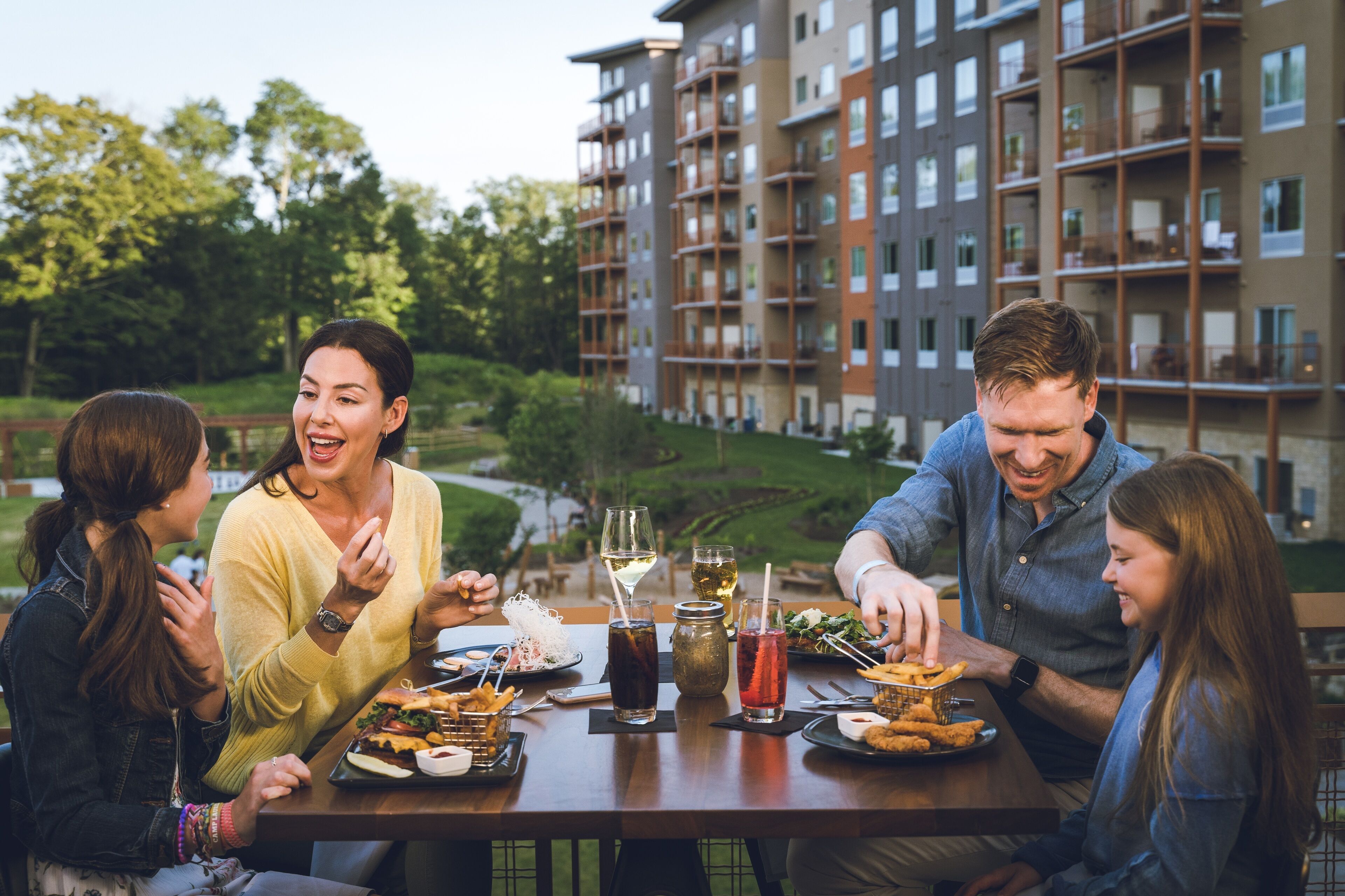 A group of people dining at an outdoor table with various dishes and drinks, set against a backdrop of a multi-story building and greenery.