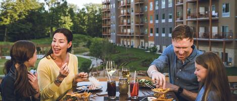 A group of people dining at an outdoor table with various dishes and drinks, set against a backdrop of a multi-story building and greenery.