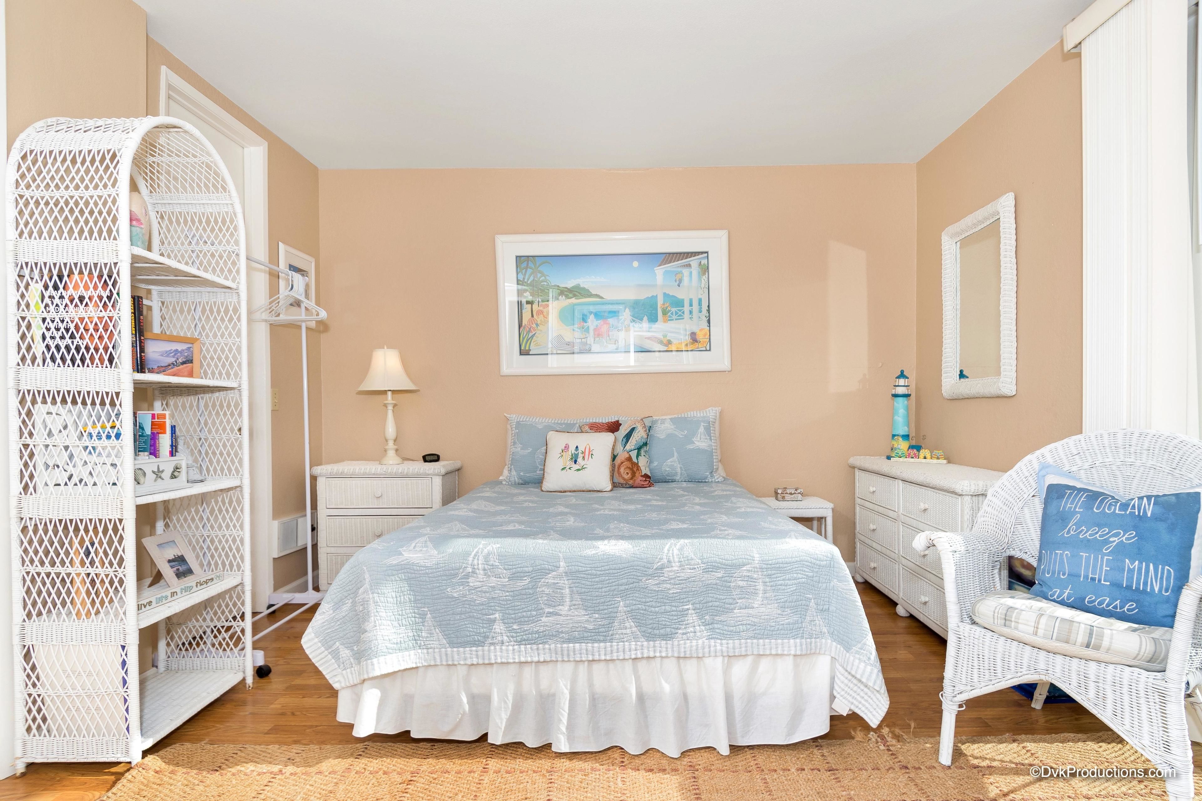 A bedroom with a bed, a white wicker side table, a white chair, a white wardrobe, and a framed picture on the wall.