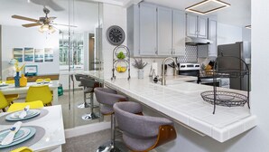A modern kitchen with white cabinetry, a large island with bar stools, and a dining area with yellow chairs. - Surfside Life (Oceanside)