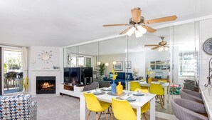 A modern living room with a fireplace, dining area with yellow chairs, and a kitchen visible through a glass partition. - Surfside Life (Oceanside)