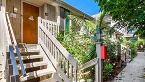 A residential building with a wooden front door, a staircase with white railings, and a small garden area with palm trees and shrubs. - Surfside Life (Oceanside)