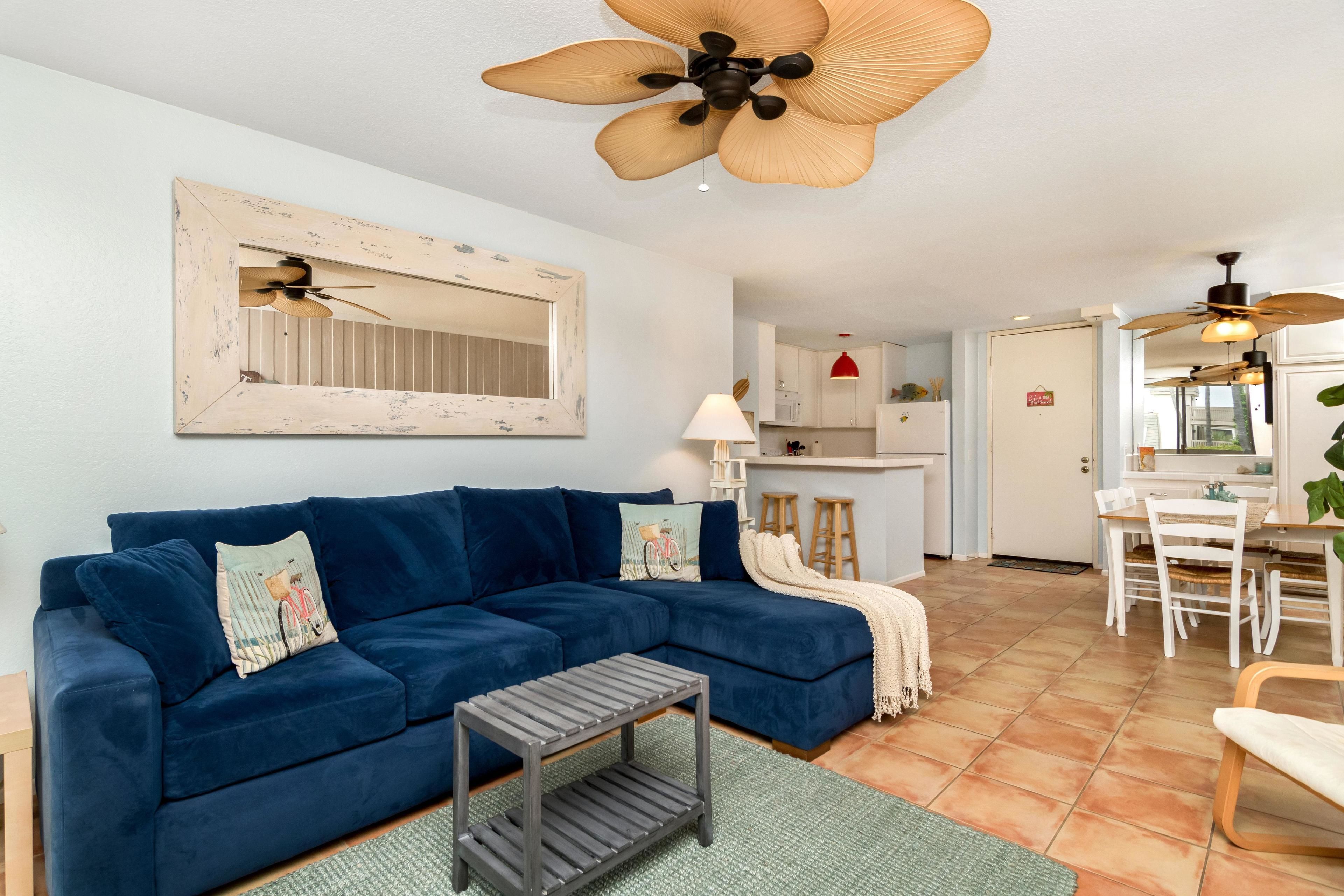 A living room with a blue sectional sofa, a small coffee table, and a ceiling fan. There is a kitchen area with white cabinets and a red hanging light.