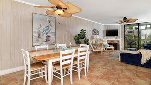 A spacious living area with a wooden dining table, white chairs, and a blue sofa. There are ceiling fans, a TV, and a fireplace. - Paradise Found (Oceanside)