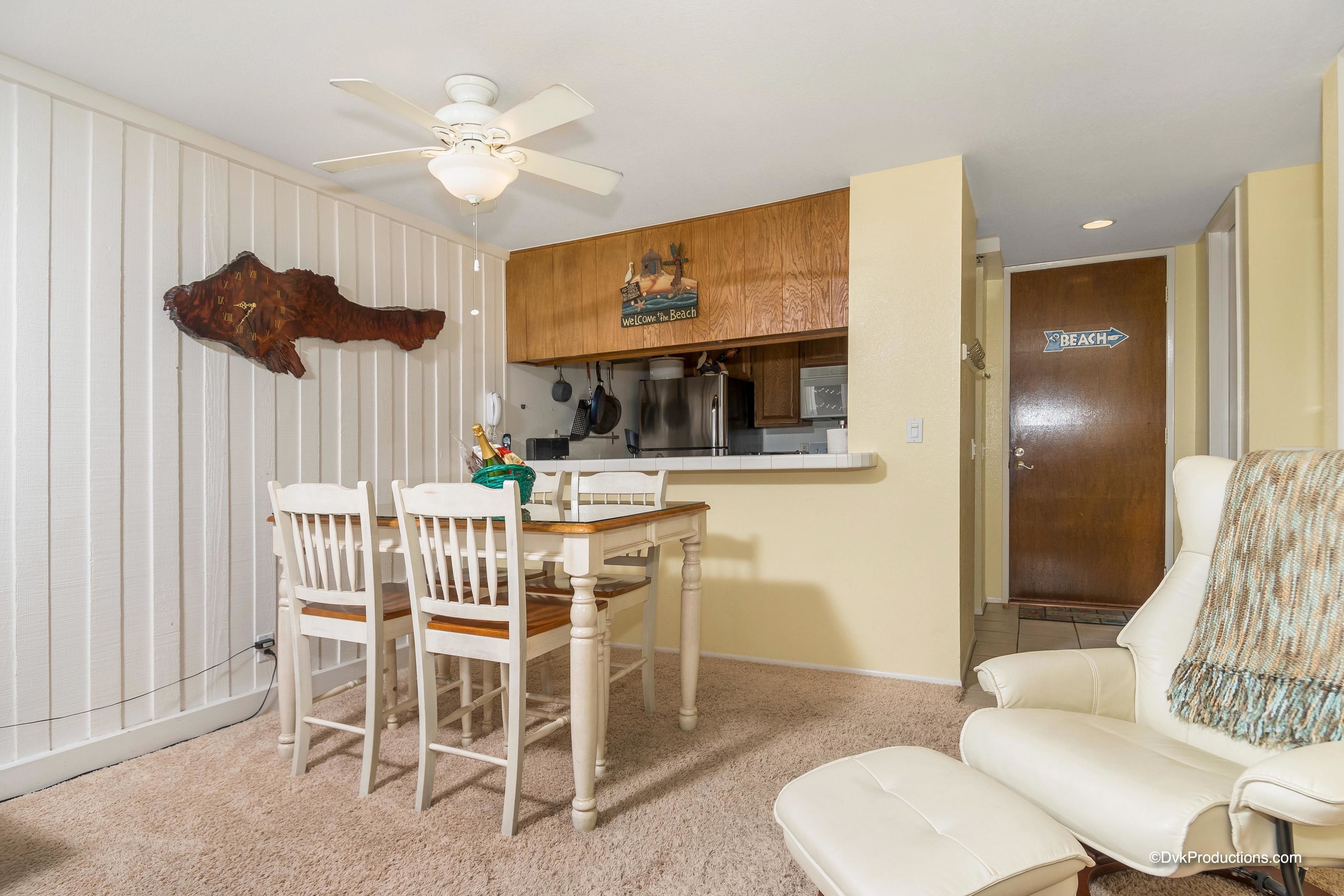 A dining area with a wooden table and chairs, a ceiling fan, and a kitchen area with wooden cabinets and a refrigerator.