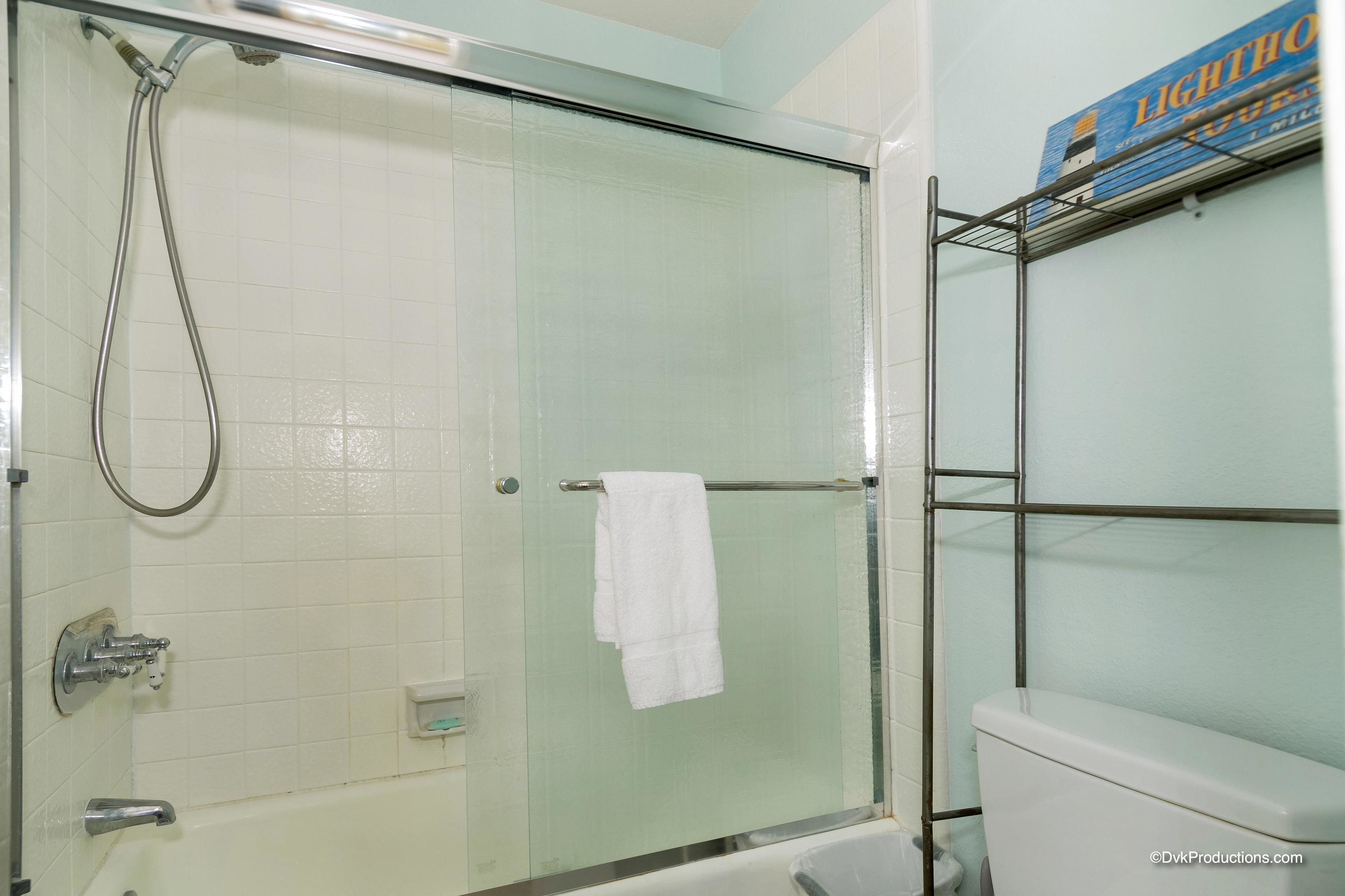 A glass-enclosed shower with a white towel hanging on the door handle, a white toilet, and a shelf with a box labeled "LIGHTHOUSE".