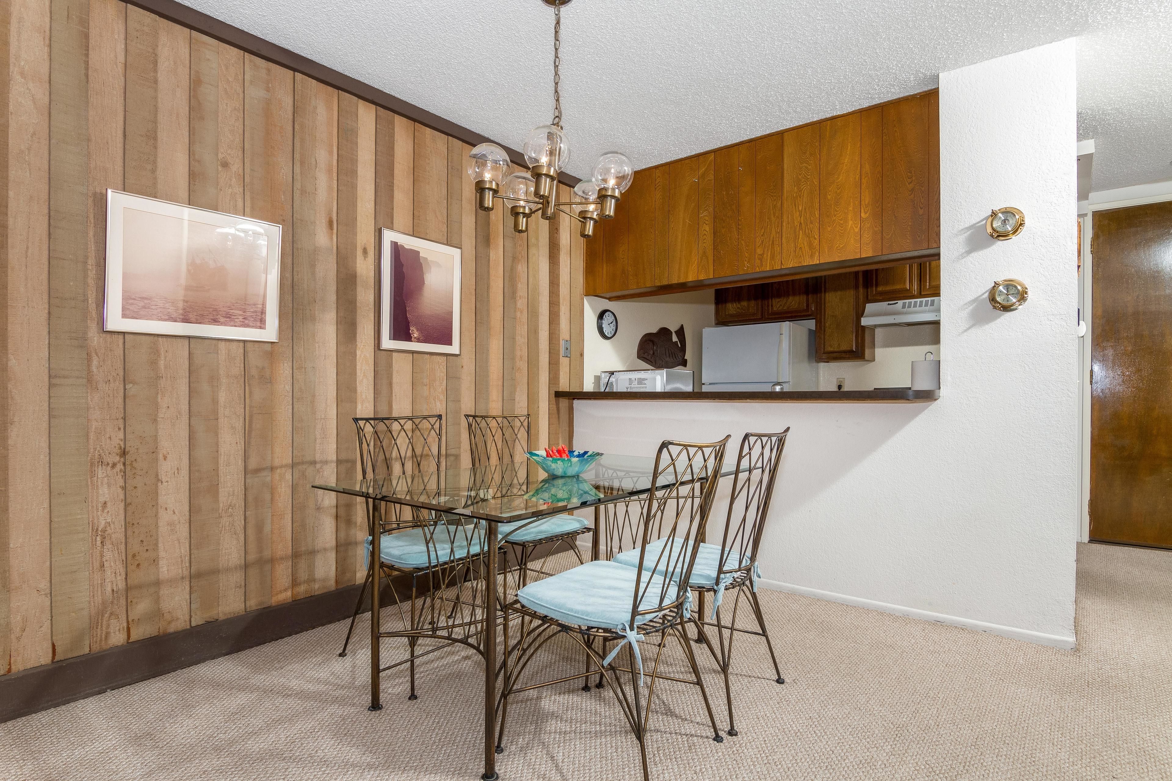 A dining area with a glass table and metal chairs, wooden panel walls, and a kitchen in the background.