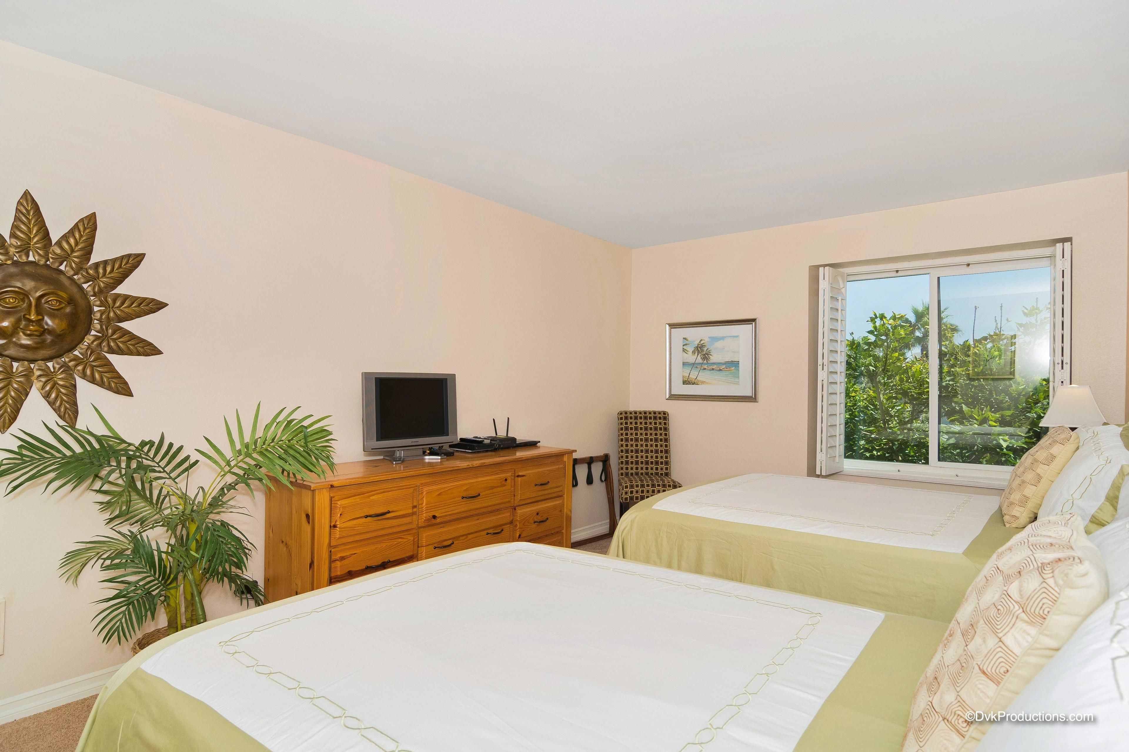 A bedroom with two beds, a wooden dresser, a television, a large sun decoration, a framed picture, and a window with a view of greenery.