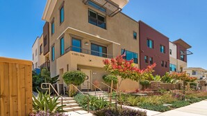 Un edificio residencial de varios pisos con fachada color beige, ventanas con marcos azules y un pequeño jardín delantero con diversas plantas y una cerca de madera.