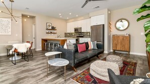 Una sala moderna con un sofá gris, un comedor con una mesa blanca, y una cocina con armarios blancos y electrodomésticos de acero inoxidable.