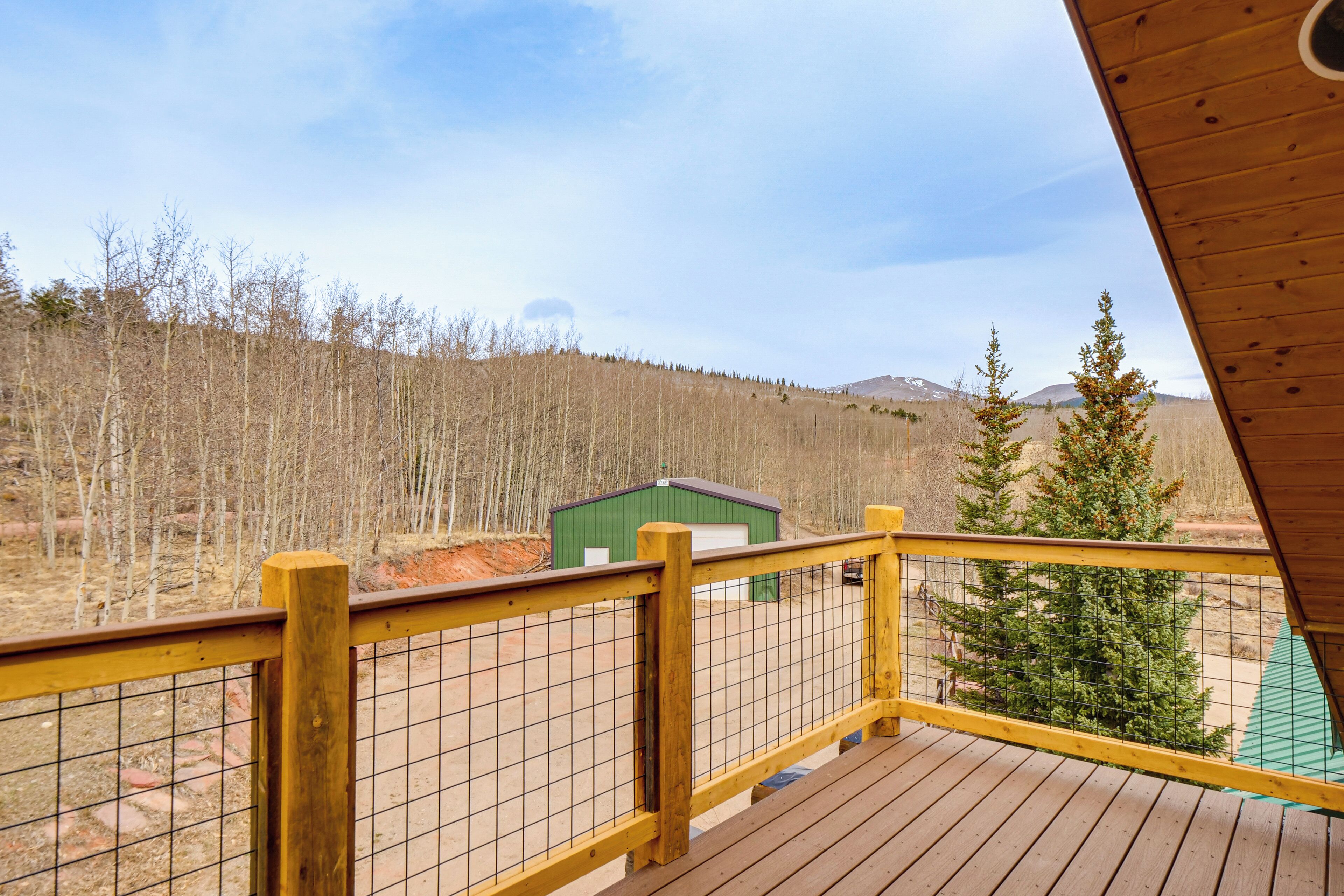 A wooden deck with a yellow railing, a green building in the background, and a forested hillside.