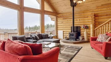 A log cabin interior with a fireplace, red sofas, a glass coffee table, and large windows offering a view of the forest.