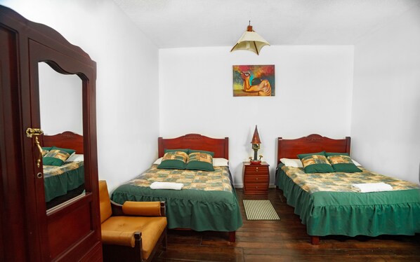 Una habitación con dos camas, una silla de madera, una mesita pequeña, una lámpara y un cuadro en la pared.