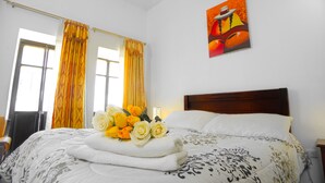 Una cama bien tendida con sábanas blancas y un edredón estampado con motivos florales, un ramo de rosas amarillas y blancas, y un cuadro de un sombrero en la pared.