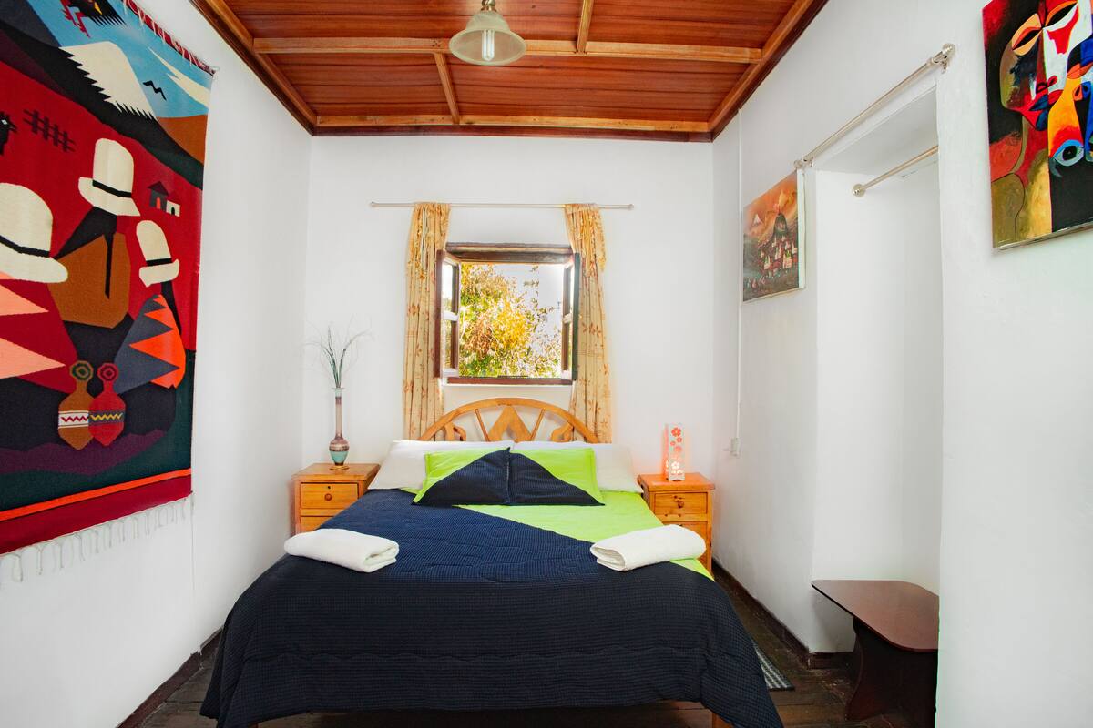 A bedroom with a bed, wooden headboard, a window with curtains, a nightstand, a bedside table with a lamp, and a wall tapestry.