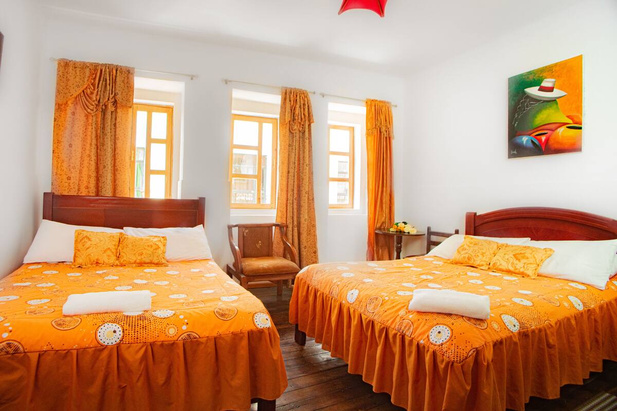 A hotel room with two beds, wooden headboards, orange bedding, a wooden chair, a small table with a vase of flowers, and a painting on the wall.