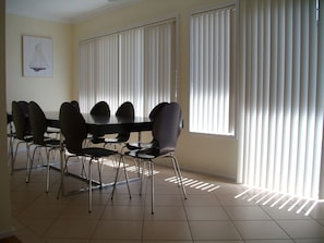 Un coin repas avec une table ronde et des chaises, de grandes fenêtres équipées de stores et un tableau représentant un voilier accroché au mur.