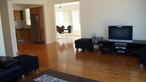 Un amplio salón con suelo de madera, un sofá negro, un televisor y una zona de comedor con sillas y una mesa.