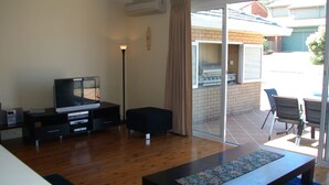 Una sala de estar con televisor, sofá, mesa de centro y puerta corrediza de vidrio que da a un patio exterior con una parrilla.